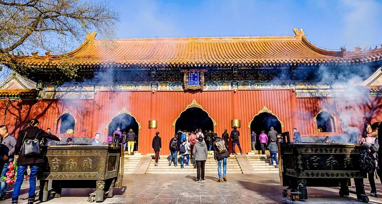 Visitors burn incense outside a red-walled temple as fragrant smoke drifts upward.