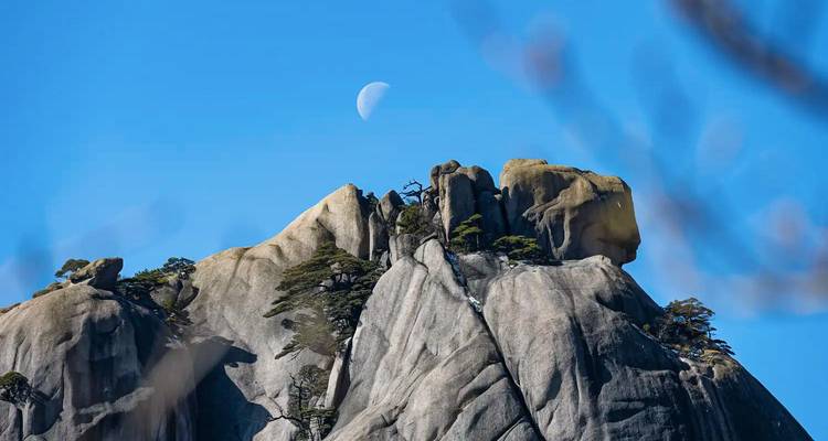 Grijze granitische pieken van Huangshan rijzen scharp af tegen een helderblauw luchtruim met een zichtbare halfmaan en door de wind geteisde dennenbomen.