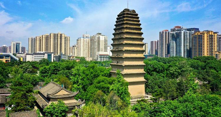 Ancient brick pagoda rises amid leafy park with modern Xi’an skyline towering behind.