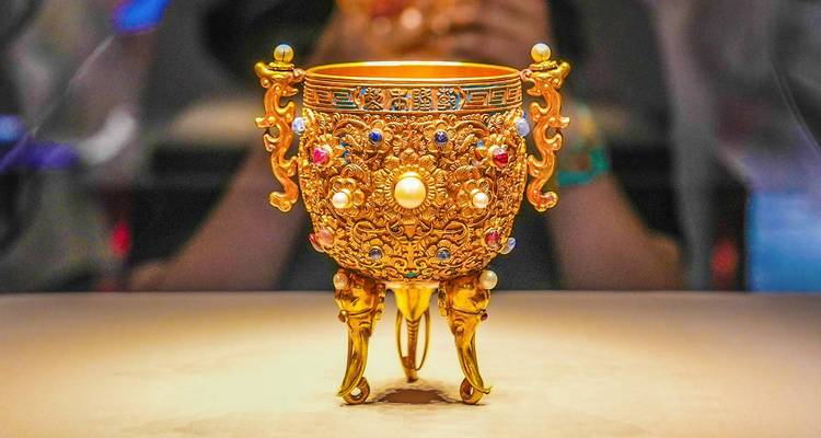 Intricately jeweled golden chalice displayed under spotlight in museum exhibit case.