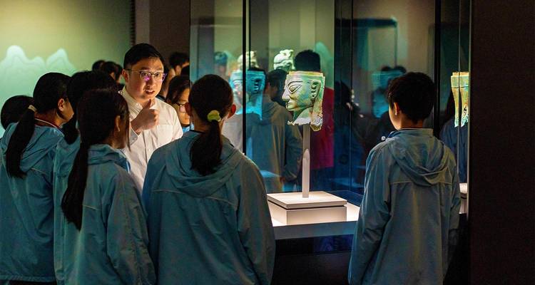 Museum guide explains an ancient bronze mask to attentive students in a dimly lit exhibit.