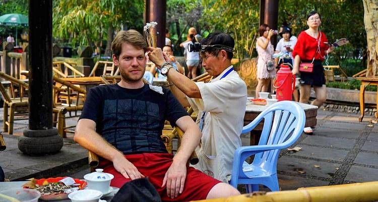 Local practitioner performs traditional ear cleaning for a visitor in an open-air tea garden.