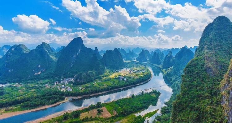 Vista aérea de picos de karst dentados alineados junto a un río sinuoso bajo un cielo azul brillante con nubes cúmulos
