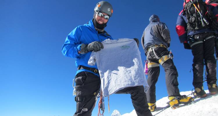 A person in climbing gear holding a shirt on a snowy peak.