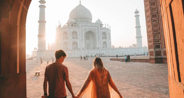 Couple hand-in-hand facing the Taj Mahal bathed in soft sunrise light through the archway.