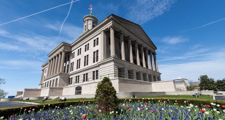 Grand neoclassical state capitol building with columns and tower set against a bright blue sky and blooming tulip garden.