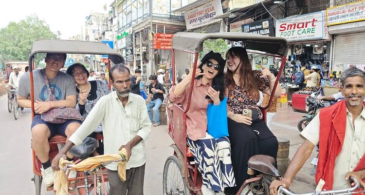 Mehrere Fahrradrikschas mit lachenden Touristen bewegen sich durch eine lebhafte indische Basarstraße.