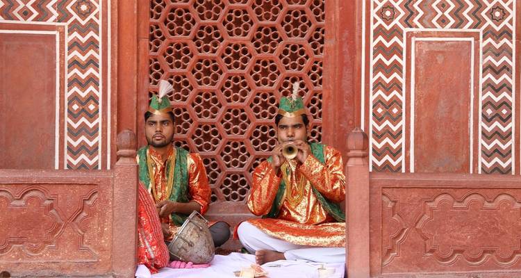 Two men in traditional attire playing instruments against a patterned wall.