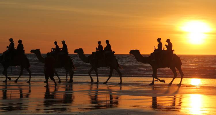 Las siluetas de jinetes en camellos cruzan una playa reflectante contra una puesta de sol naranja intenso sobre el océano.