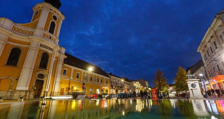 Een stadsplein in de schemering met verlichte gebouwen en een vijver die het licht weerkaatst.