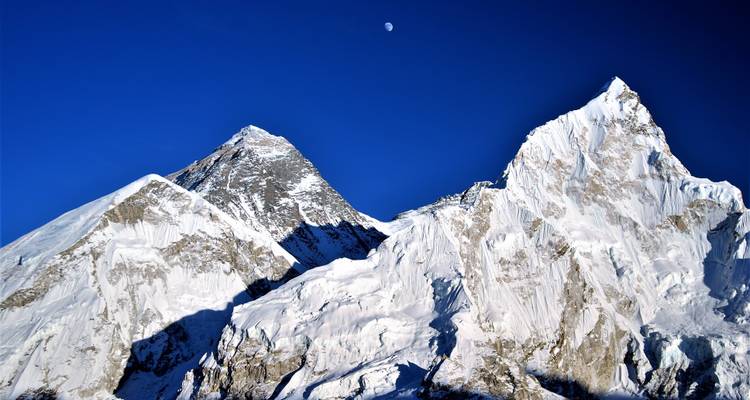 Picos de montañas cubiertas de nieve con la luna visible en el cielo azul.