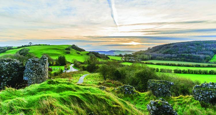 Rolling green Irish countryside dotted with ruins under a soft, streaked sunset sky.