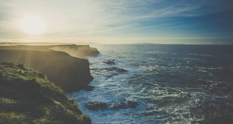 Rugged cliffs of Moher meet crashing Atlantic waves beneath hazy sunset light.