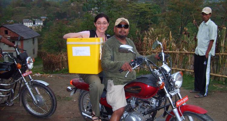 Un voyageur se tient en croupe sur une motocyclette rouge conduite par un habitant local à travers un village rural népalais.