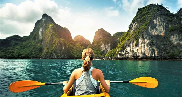 Back view of a woman paddling a yellow kayak on turquoise water surrounded by dramatic limestone karsts