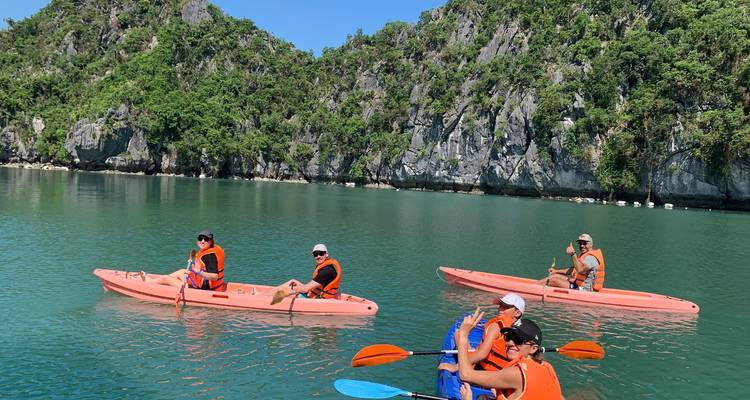 Several kayakers in orange life jackets paddle on calm emerald water beneath green limestone cliffs