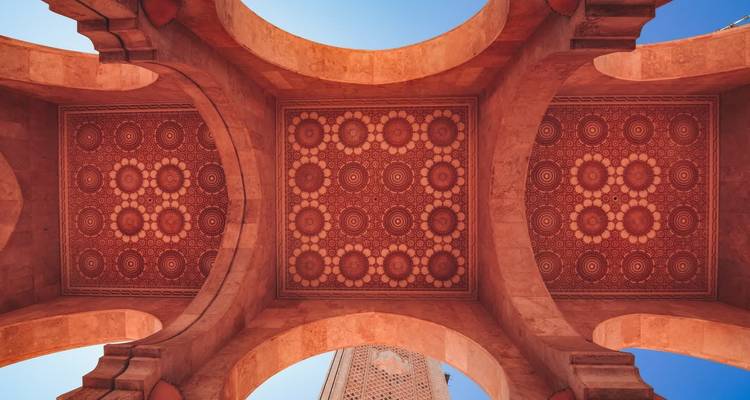 Symmetrical view of ornate red patterned ceiling and stone arches of a Moroccan mosque with blue sky beyond.