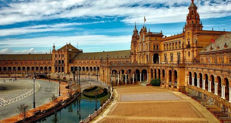 Wide panorama of the red-brick Plaza de España building in Seville curving around a canal under a bright blue sky.