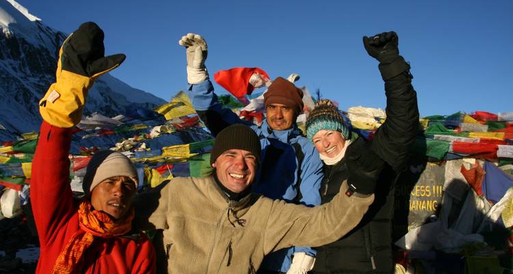 Happy trekkers celebrating with raised arms beside fluttering prayer flags against high snowy peaks