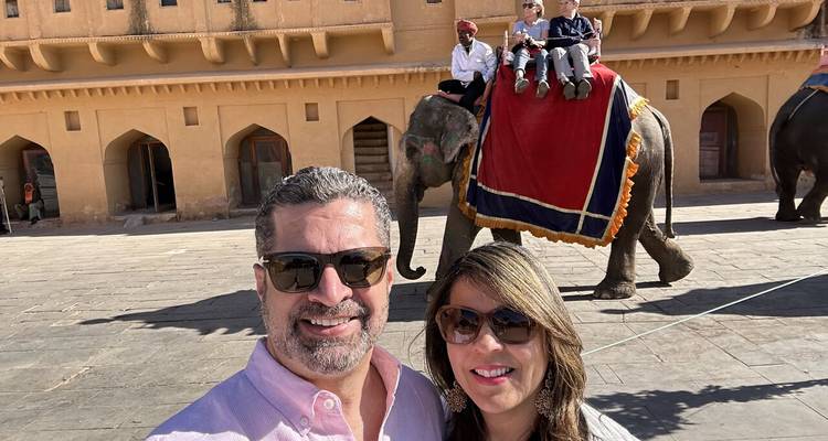 Two people posing in front of a historic building with an elephant carrying riders in the background.