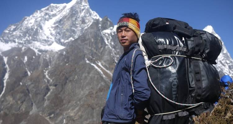 Een sherpa-drager draagt een groot zwart rugzak langs een bergpad met sneeuwtoppen die dreigend opdoemen op de achtergrond.