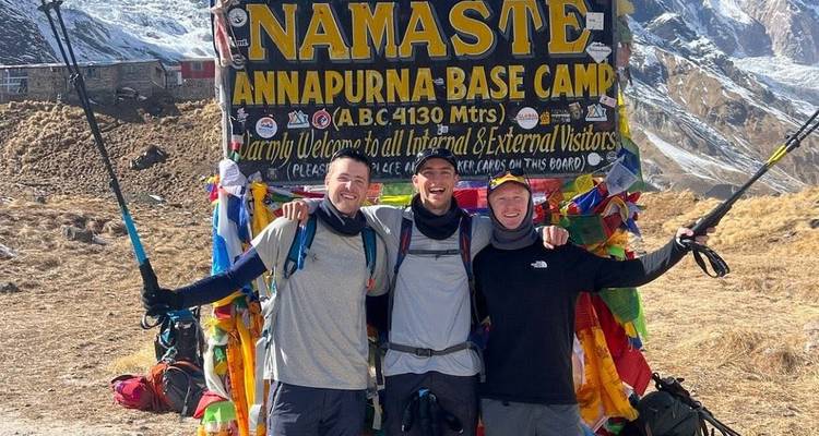 Drie trekkers glimlachen arm in arm onder het kleurrijke welkomstbord van Annapurna Base Camp.