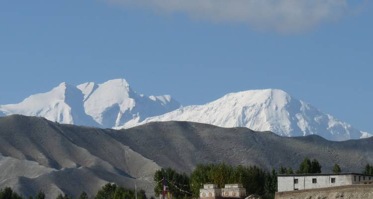 Sneeuwbedekte Himalaya-pieken torenen uit boven de kale grijze heuvels en een klein nederzettinkje eronder.