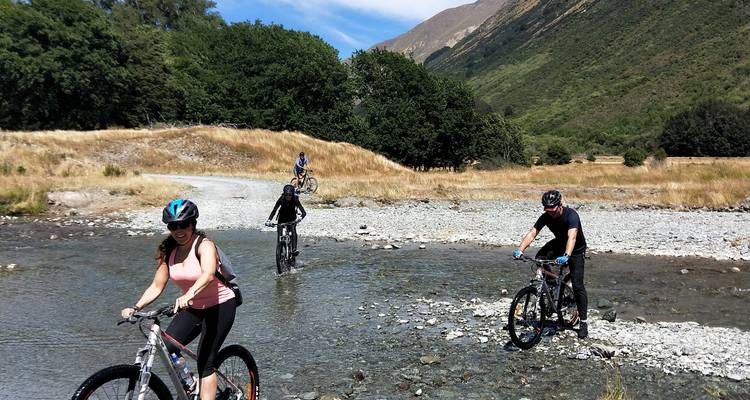 Des cyclistes traversent une rivière peu profonde entourée de collines.
