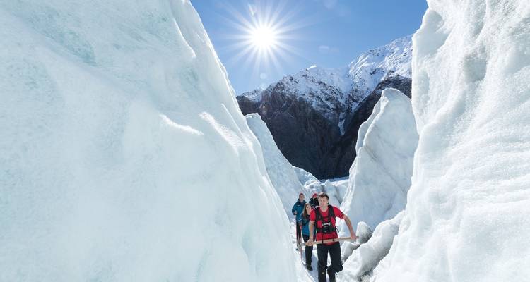 Menschen, die durch eisige Spalten wandern mit Blick auf schneebedeckte Berge unter einer strahlenden Sonne.