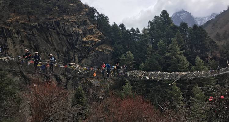 Pont suspendu avec des randonneurs et des drapeaux de prière au-dessus d'une vallée boisée.