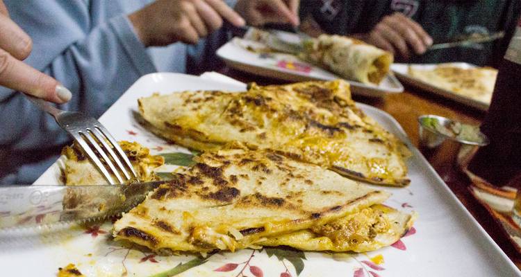 Des mains découpent des quesadillas au fromage sur un plateau à motifs floraux lors d'un repas décontracté.