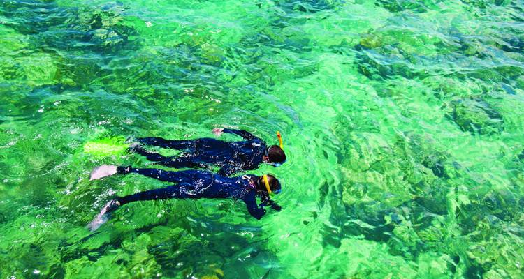 Two snorkelers glide over crystal-clear emerald water filled with coral formations.