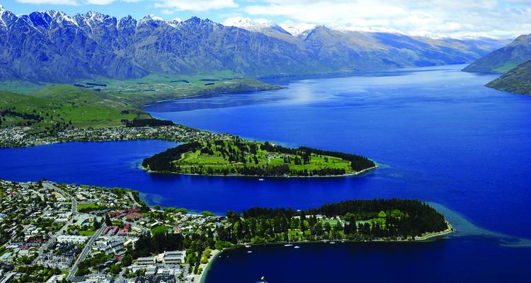 Aerial vista of Queenstown with a blue lake, pine-covered peninsula and surrounding snow-capped mountains.
