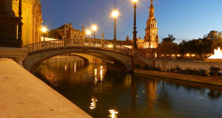 Puente y torre iluminados de la Plaza de España reflejándose en el canal al atardecer.