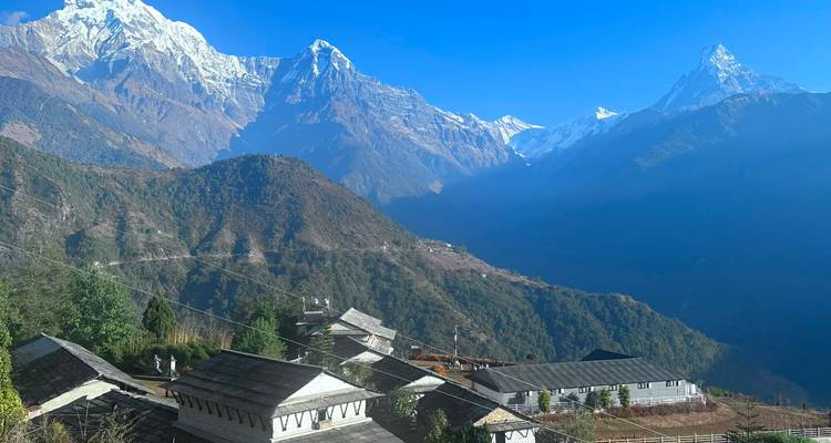 Les toits des maisons d'un village de montagne se dressent face à une vallée d'un vert profond avec des pics enneigés imposants sous un ciel bleu dégagé.