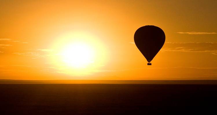 La silueta de un globo aerostático se desliza contra un cielo naranja brillante al amanecer sobre la llanura infinita