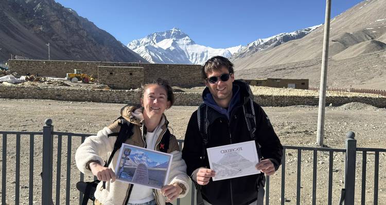 Dos montañeros sosteniendo certificados del Monte Everest con el Everest cubierto de nieve asomando detrás