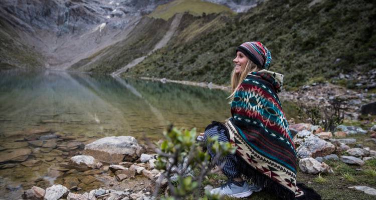 Voyageur souriant enveloppé dans un poncho coloré assis au bord d'un lac alpin tranquille avec des pics escarpés.