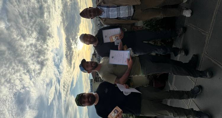 Cuatro hombres sosteniendo plantas y certificados con un fondo de atardecer.