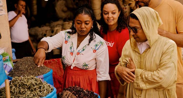Des voyageurs examinent des tas colorés d'épices dans des sacs à un étal animé de souk marocain.