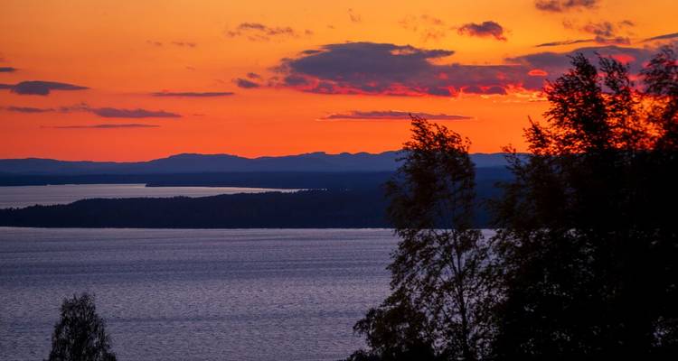 Leuchtend orangener Sonnenuntergang über ruhigen nordischen Gewässern mit silhouettierten Uferlandschaften.
