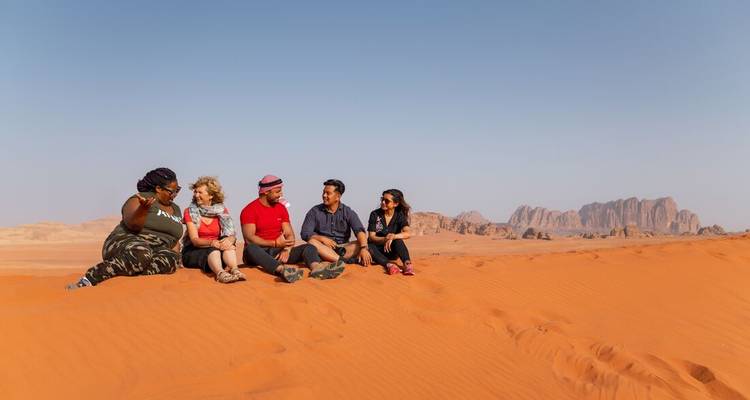 Groep die op rode zandduin zit te praten met rotsformaties van Wadi Rum op de achtergrond