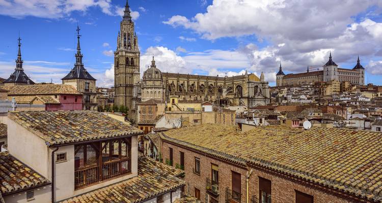 Historische Skyline von Toledo, dominiert vom aufragenden Kathedralenturm und Alcázar über Terrakotta-Dächern unter einem strahlend blauen Himmel.
