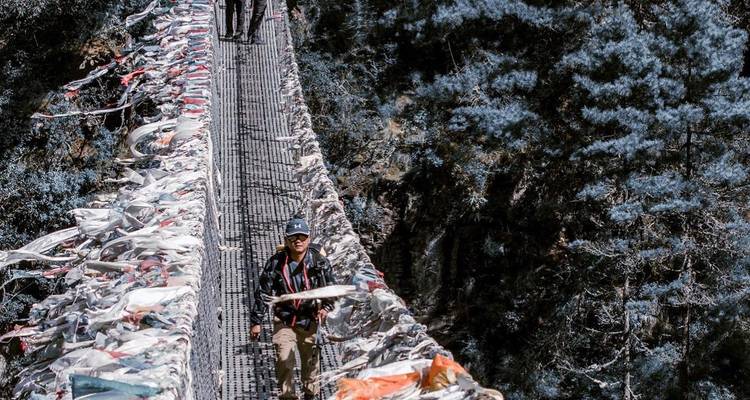 Una persona caminando sobre un puente colgante decorado con banderas de oración.