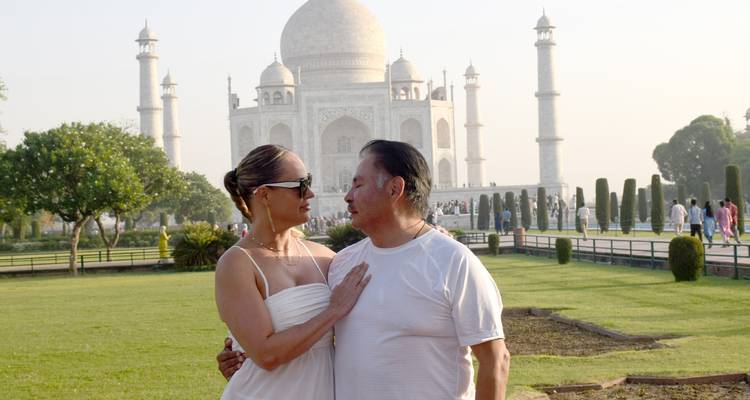Couple posing affectionately in front of the Taj Mahal in hazy morning light, green lawns in foreground.