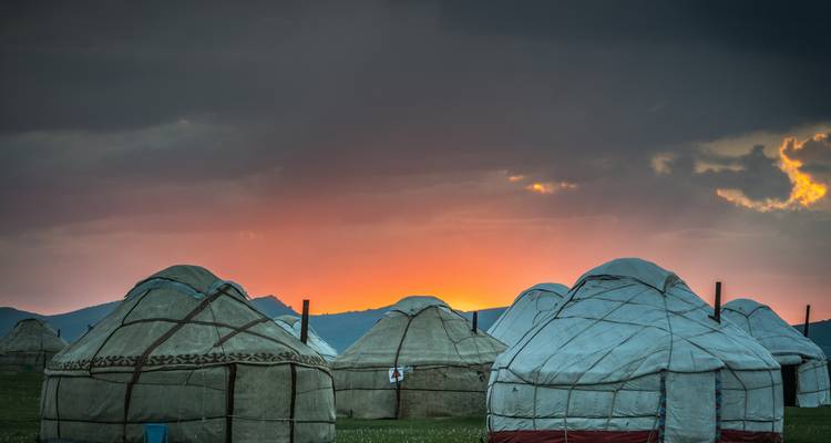 Row of yurts silhouetted against a vivid orange sunset on an open high-altitude plain.