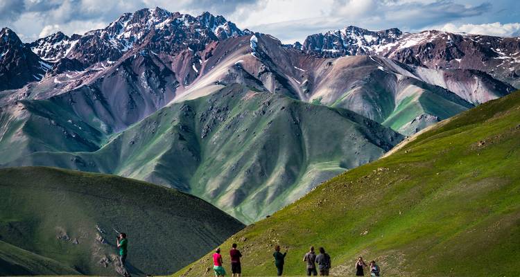 Small group of hikers admire rolling green valleys and rugged snow-capped peaks.