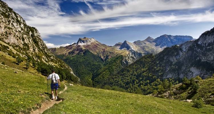 Ein Wanderer wandert auf einem bewachsenen Bergrücken entlang, mit dramatischen Pyrenäengipfeln und gestreiften Wolken in der Ferne.