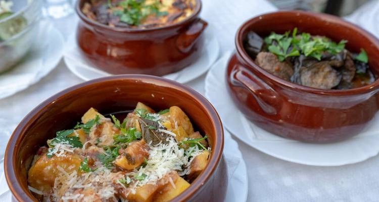 Primer plano de tres cuencos de loza llenos de guisos tradicionales abundantes y pastas en una mesa de comedor al aire libre.
