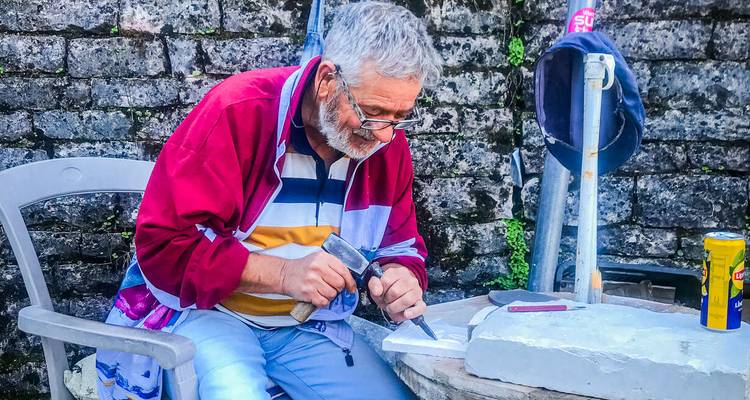 Un artesano anciano en una chaqueta colorida cincela piedra en un pequeño taller al aire libre contra una pared de ladrillo rugosa.